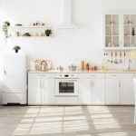 Kitchen with light walls, white furniture and shelves with crockery and plants in pots, small refrigerator in dining room scandinavian design, empty space. Modern interior with sunbeams on the floor